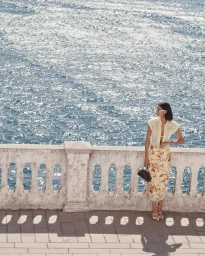 Model wearing patterned midi skirt and blouse leaning against a stone balustrade overlooking a sparkling ocean by Benny Horne
