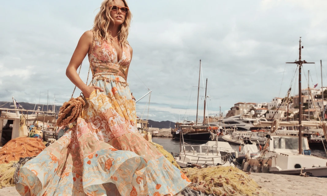 Woman in patterned dress posing on dock with harbor, boats, and town in background under cloudy sky. Fashion editorial, travel photography.