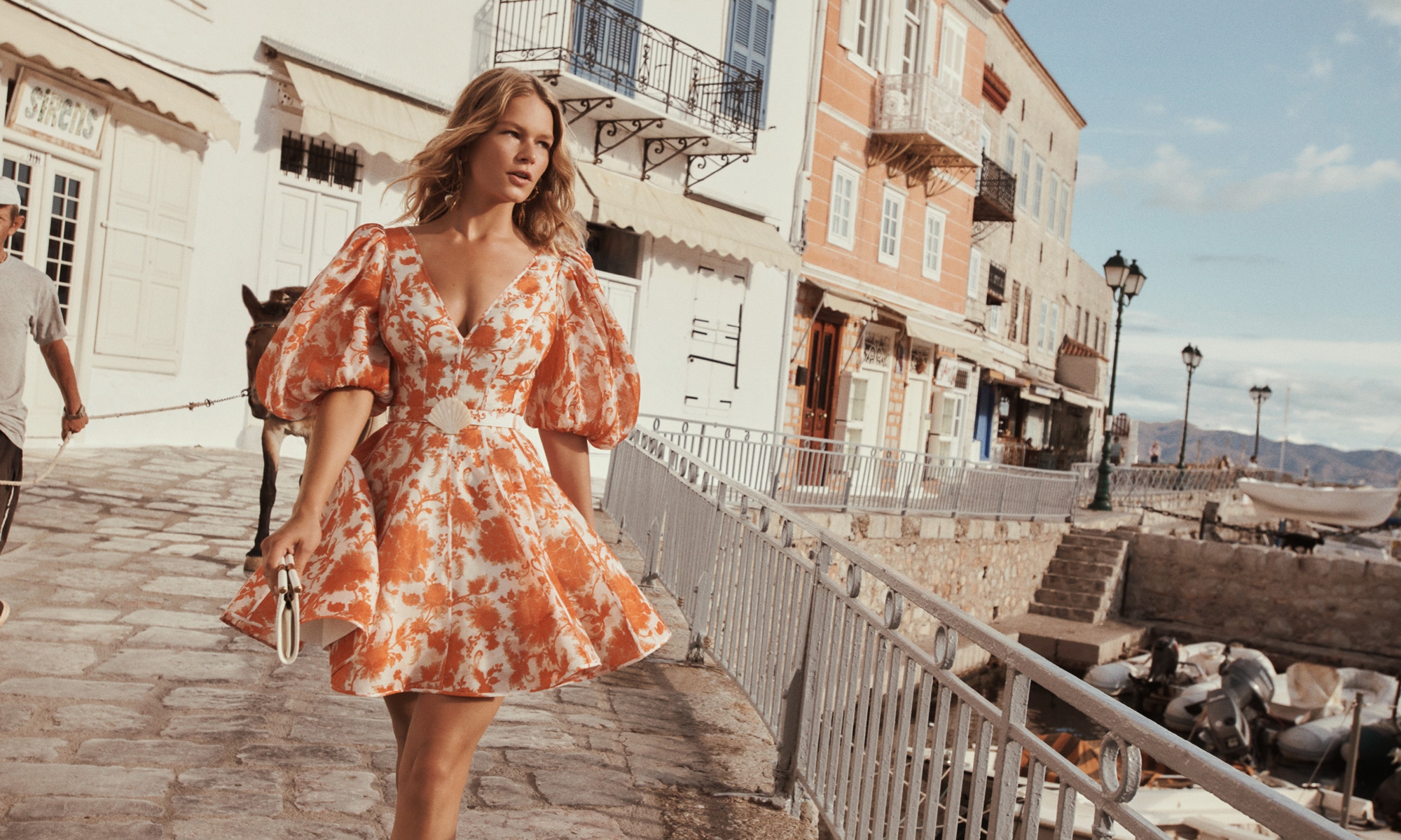 A blonde woman in a short orange-and-white floral dress walks along a stone promenade with a donkey in a seaside village.