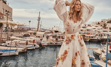 Model Anna Ewers in a floral applique white dress posing by a harbor filled with boats for Zimmermann Resort RTW.