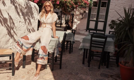 Model Anna Ewers sitting on a cafe table wearing a printed cream jumpsuit for Zimmermann Resort RTW.