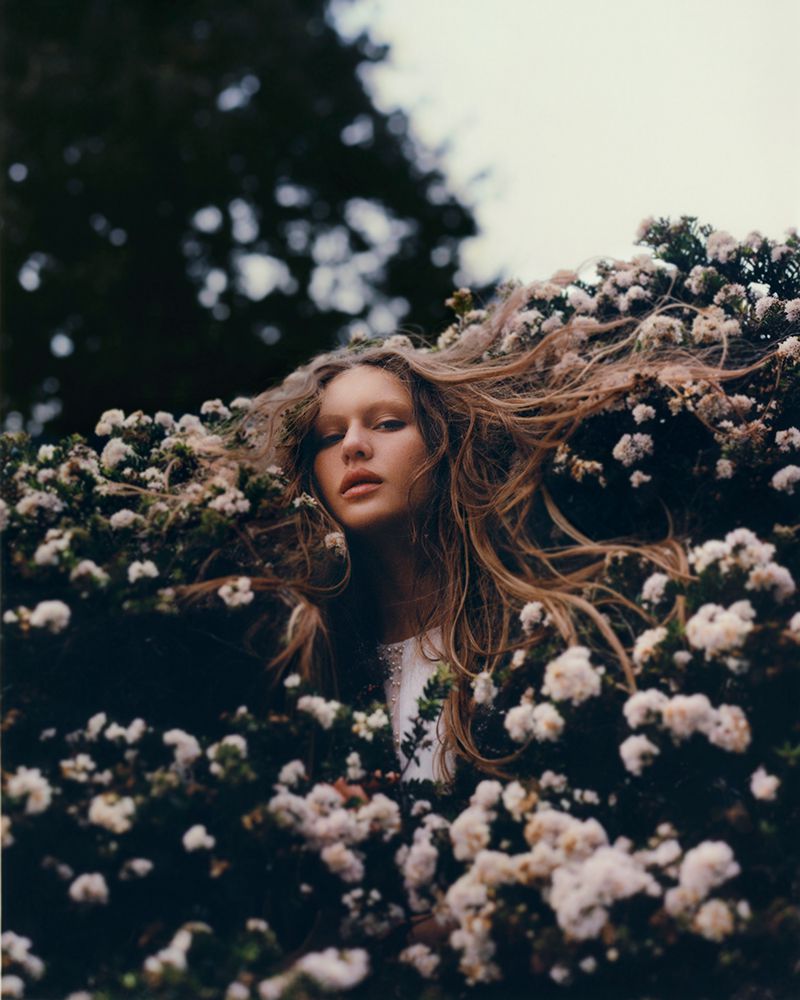 Portrait of a fair-skinned young woman with long blond hair surrounded by flowering branches, gazing into the camera in outdoor setting.