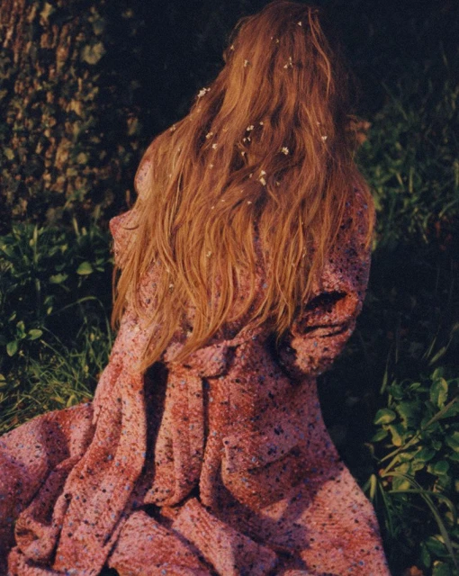Back view of an adult female with long red hair, wearing a patterned pink jacket, standing in a grassy outdoor setting with tree shadows.