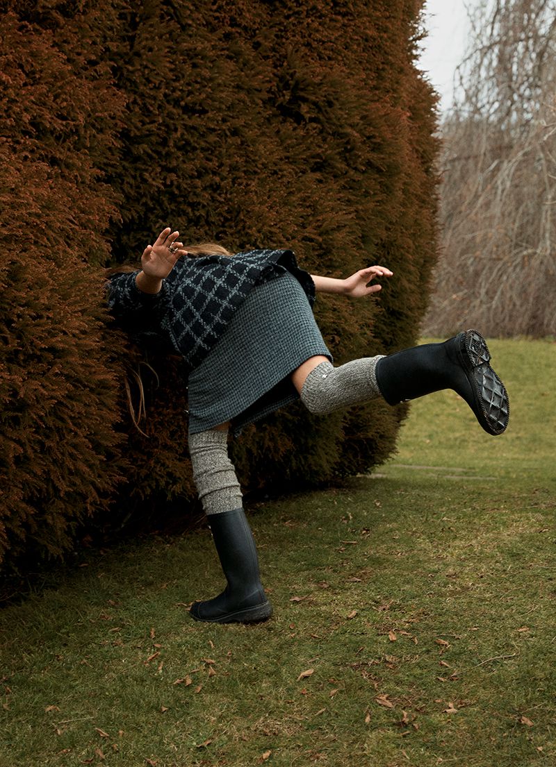 A single person is seen playfully entering a large, rounded hedge in an outdoor grassy area, wearing a patterned sweater, skirt, and boots.