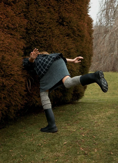 A single person is seen playfully entering a large, rounded hedge in an outdoor grassy area, wearing a patterned sweater, skirt, and boots.