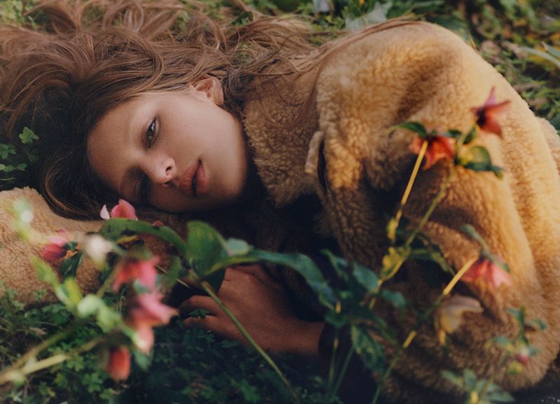 A young adult female model with brown hair lies on a grassy field surrounded by small pink flowers wearing a sherpa coat looking at the camera.