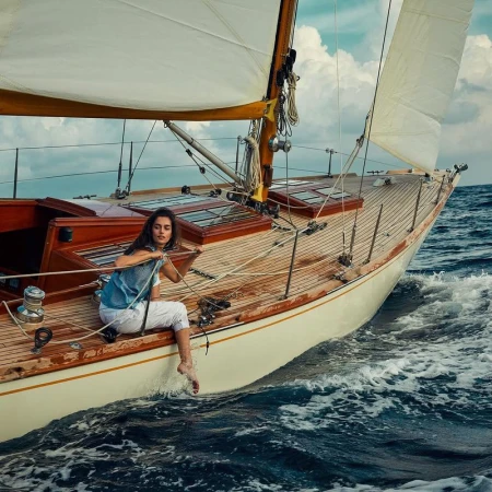 Woman sitting on the edge of a vintage sailboat, enjoying the ocean breeze. The boat cuts through the waves on a sunny day.