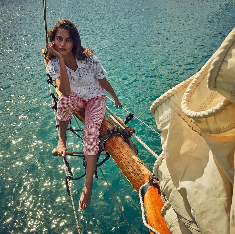 A woman in a white top and checkered pants is sitting on the bowsprit of a sailboat with sparkling turquoise water behind her.