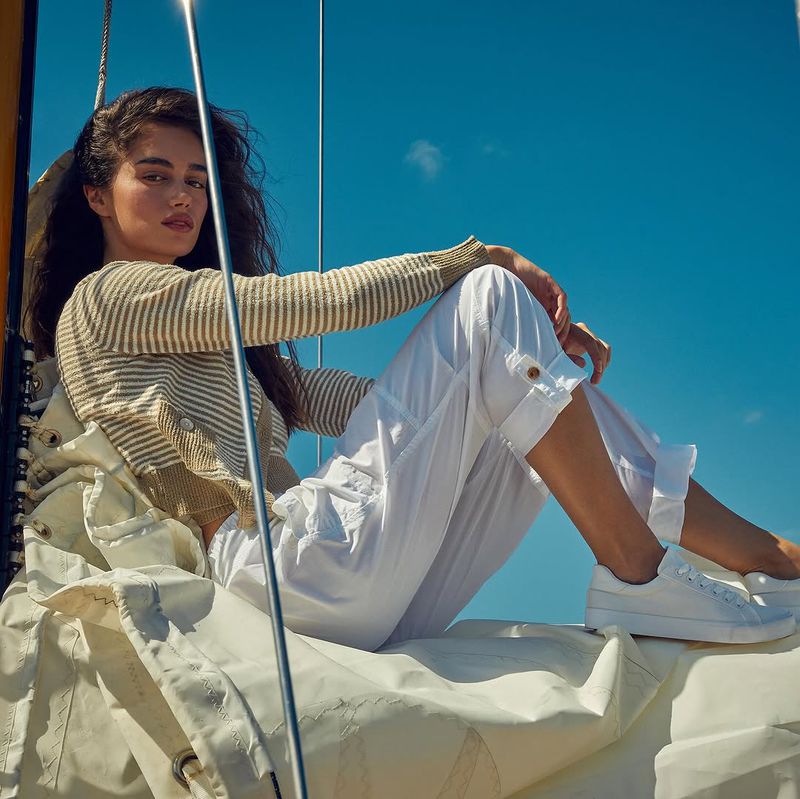 Brunette woman wearing white pants and striped sweater sits casually on a boat, enjoying the clear blue sky and sunny weather.