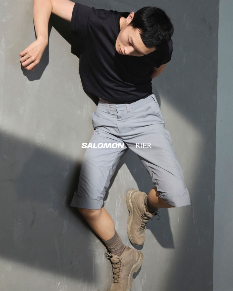 A young adult male in casual attire is lying on a grey surface next to a lighter wall, wearing a black t-shirt, grey shorts, and boots.