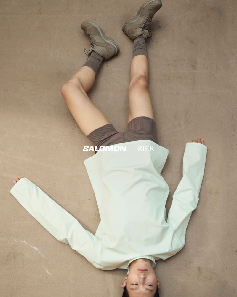 Person lying on a light brown surface, wearing a light green jacket, brown shorts and socks, and hiking shoes, presenting Salomon brand.