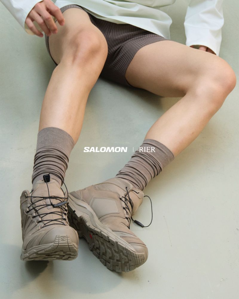 Close-up studio shot of a person wearing Salomon hiking boots with ribbed socks and shorts against a light-colored studio backdrop.
