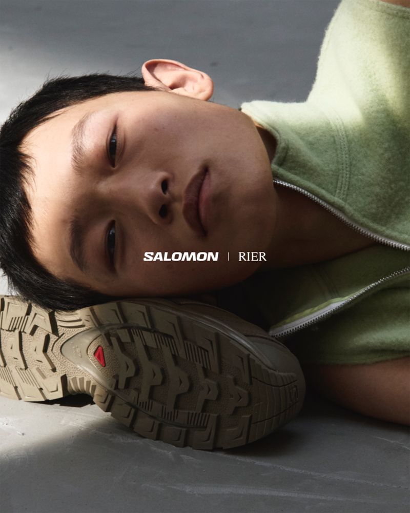 Close-up studio shot of a young Asian male model resting his head on a Salomon sneaker while wearing a green fleece top.