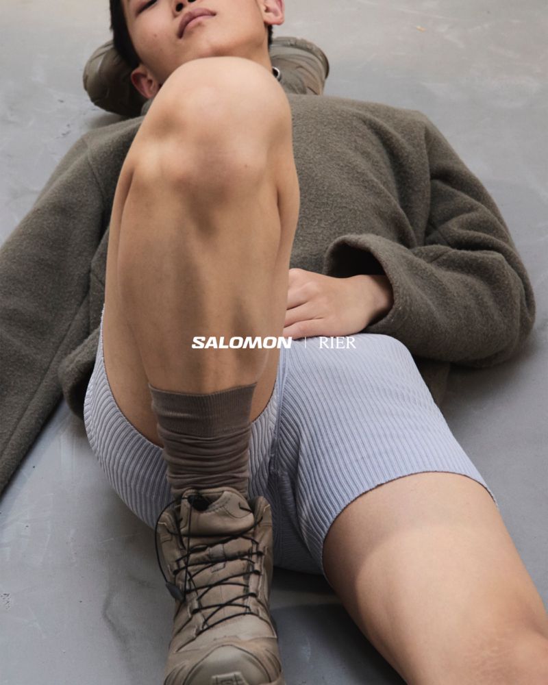 An adult male fashion model is reclining on a gray surface, showcasing grey hiking boots and socks paired with periwinkle ribbed shorts.