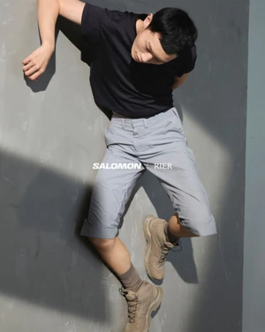 Man wearing a black t-shirt, grey shorts, and beige boots lying on a grey studio floor for RIER x SALOMON 2025 campaign.