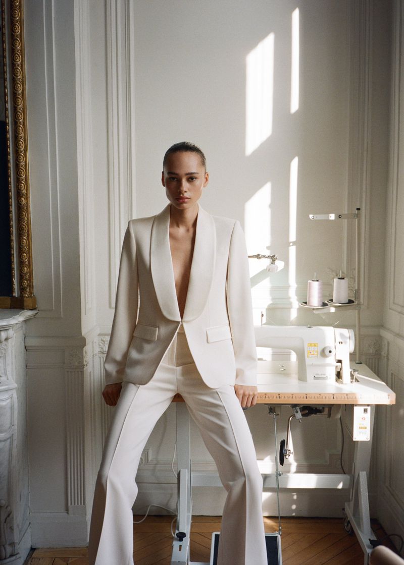 A woman in a cream-colored pantsuit poses in front of a sewing machine and white walls with light coming through a window.