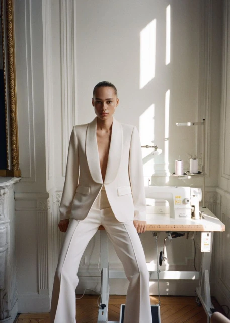 A woman in a cream-colored pantsuit poses in front of a sewing machine and white walls with light coming through a window.