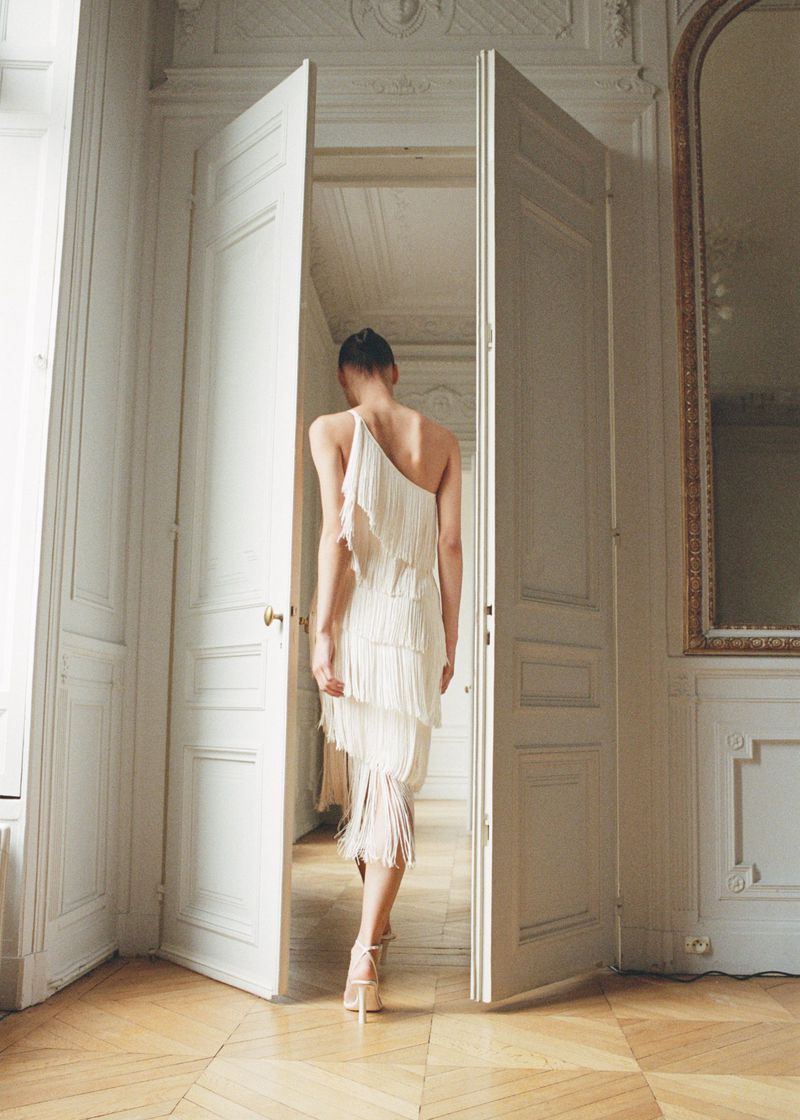 Elegant woman in a white fringe dress walks through open white doors into a classically decorated room with parquet floor.