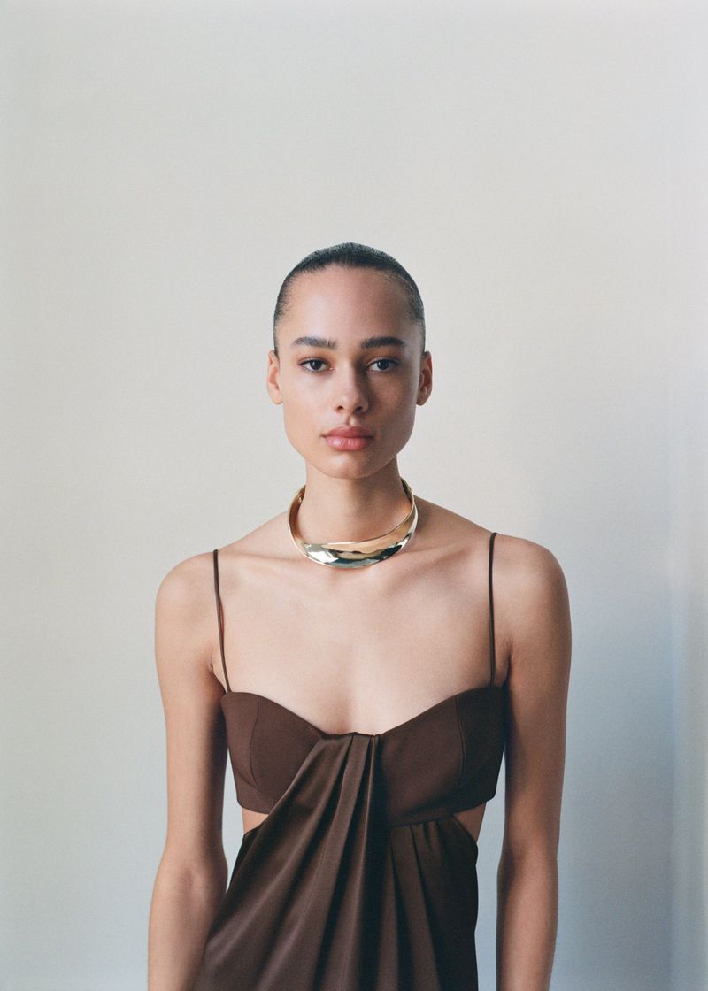 A young adult woman with a slicked-back hairstyle and a gold collar necklace poses in a brown draped dress against an off-white wall.