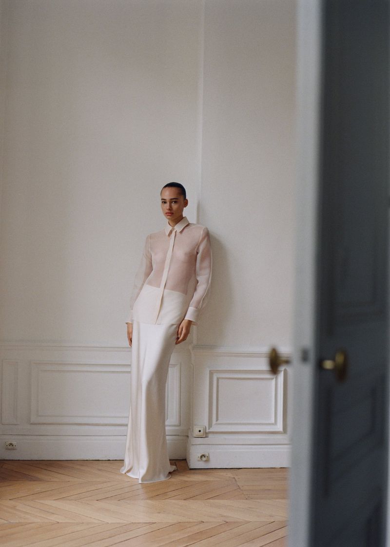 Model wearing a sheer blouse and long skirt standing against a white wall in an apartment