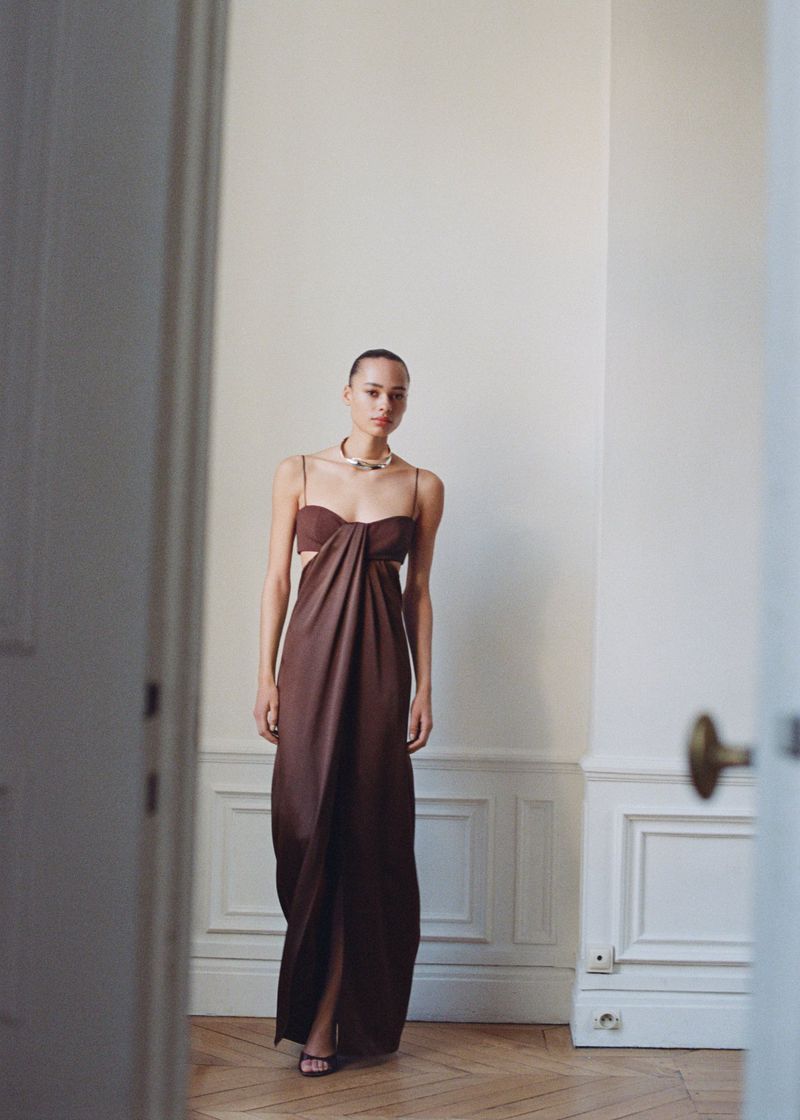 Woman in long brown cut-out dress poses indoors, framed by open doorway showing parquet flooring and cream walls with molding.