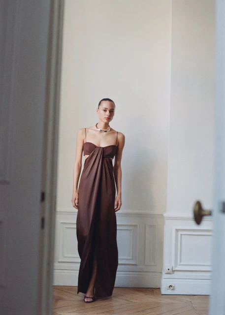 Woman in long brown cut-out dress poses indoors, framed by open doorway showing parquet flooring and cream walls with molding.