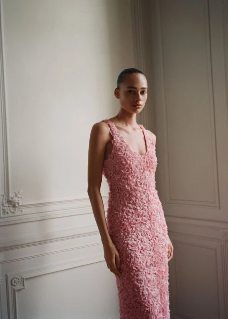 Woman wearing a pink floral textured gown stands in front of a paneled wall. Fashion shot in indoor environment.