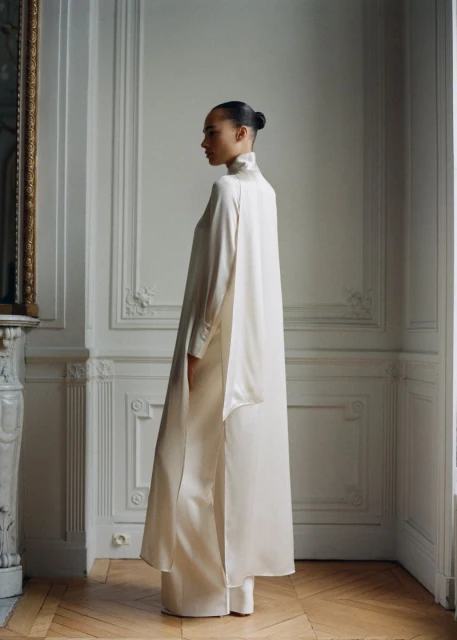 Elegant adult female wearing long-sleeve dress and trousers in a photo studio with hardwood flooring and architectural details.