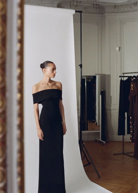 Elegant woman in a black off-the-shoulder gown posing against a white backdrop in a studio setting, part of a fashion editorial.