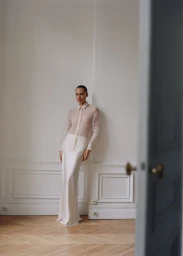 Model wearing a sheer blouse and long skirt standing against a white wall in an apartment