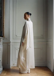 Model wearing a white satin tunic and trousers standing in a classic room with moulding and a parquet floor.