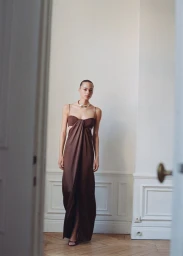 Model in a brown satin slip dress standing in an elegant interior with white walls and wood flooring.