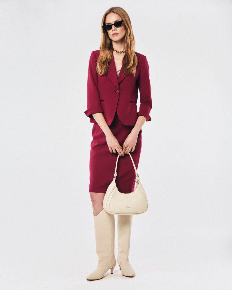 Elegant woman poses in a dark pink skirt suit, knee-high boots, and sunglasses, holding a light-colored handbag against a white background.