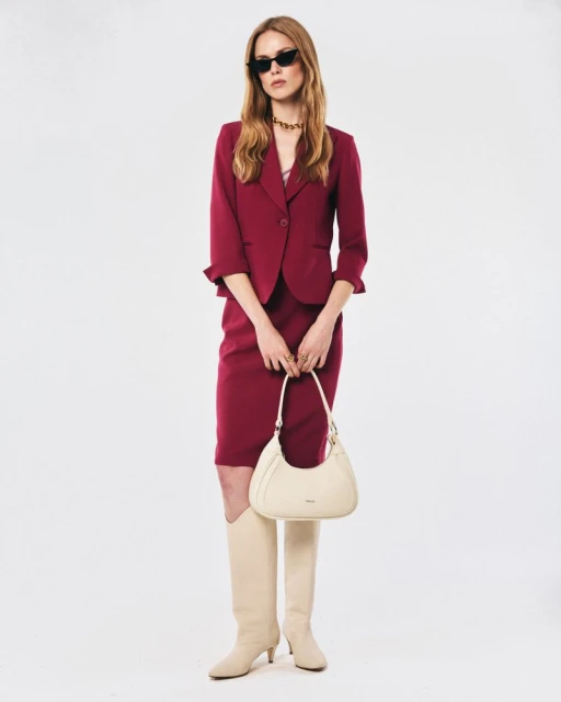 Elegant woman poses in a dark pink skirt suit, knee-high boots, and sunglasses, holding a light-colored handbag against a white background.