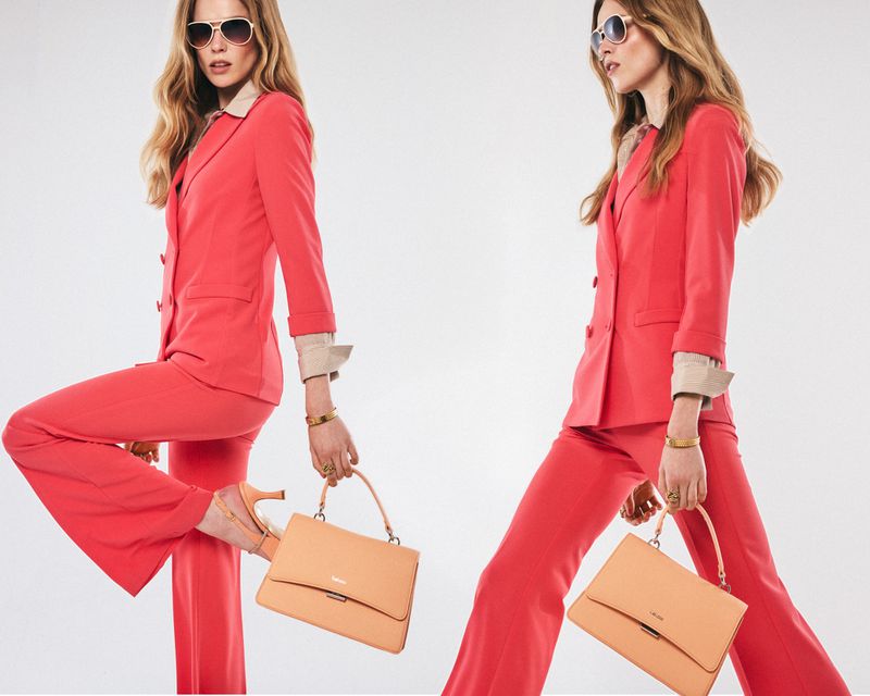 Two blonde women in red suits and sunglasses pose with handbags in a studio shot against a white backdrop, fashion-forward.