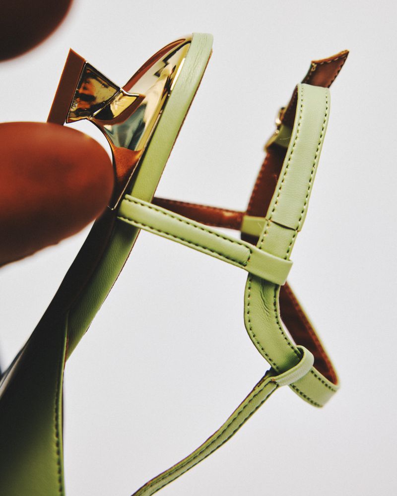 Close-up view of a light green leather heeled sandal, accentuating its intricate strap details and the shiny metallic gold heel.
