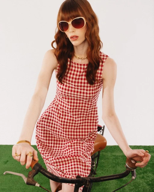 Woman in red-and-white gingham dress posing on bicycle with sunglasses against white background in a fashion studio shot. 