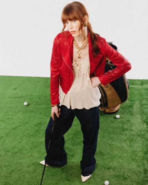 A stylish woman in a red jacket poses with a golf club on artificial turf in a fashion editorial shot, golf bag in background.