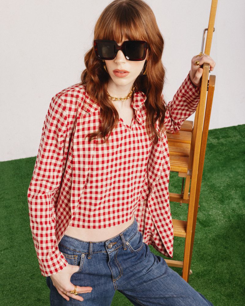 A fair-skinned young adult woman models a red-and-white gingham top and blue jeans near a wooden stepladder on a grassy indoor surface.