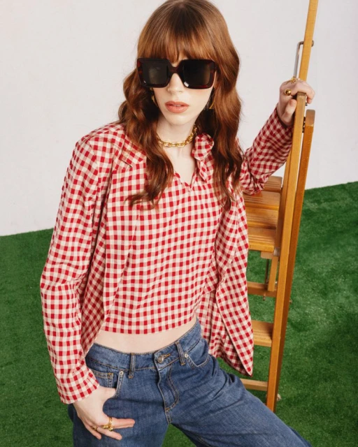 A fair-skinned young adult woman models a red-and-white gingham top and blue jeans near a wooden stepladder on a grassy indoor surface.
