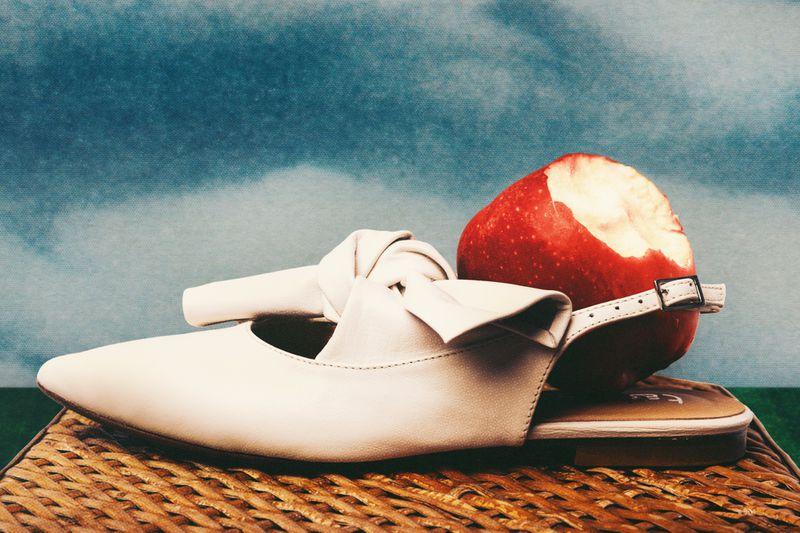 Studio shot of a white leather shoe, knotted detail, next to a red apple, on a woven surface against a textured blue backdrop.