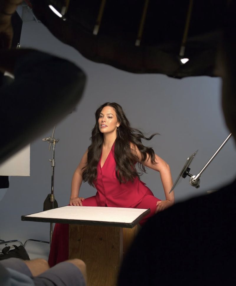 A woman in a red jumpsuit poses at a table during a photo shoot in a studio, surrounded by lighting equipment and a camera operator.
