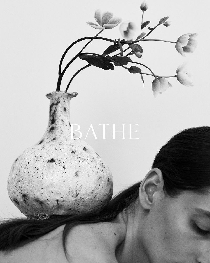 Monochromatic photograph of a woman with a vase of flowers on her back against a stark white background, creating a surreal, artistic portrait.
