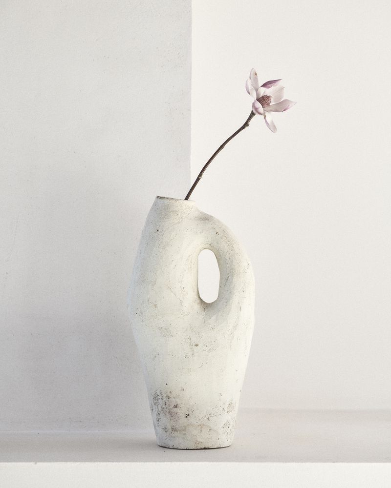 Still life of a white ceramic vase with an opening, holding a single pink magnolia blossom in a bright, minimalist interior setting.