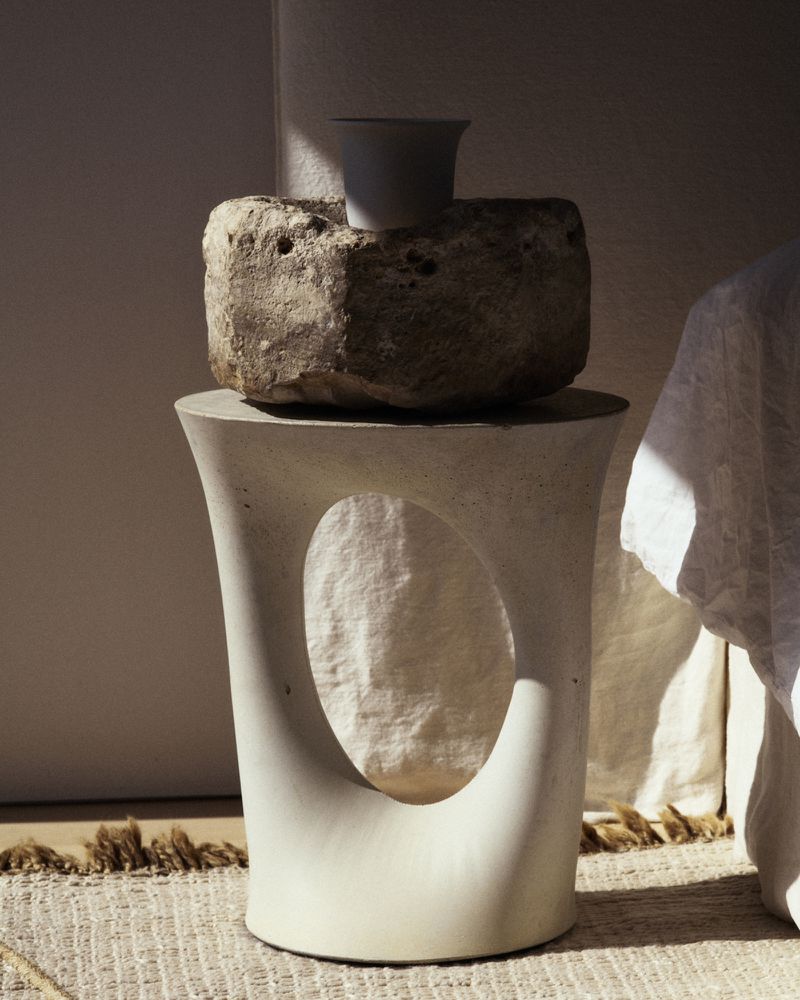 Minimalist interior still life featuring a stone block topped with a white vase, placed on a sculptural pedestal, set against a textured rug.