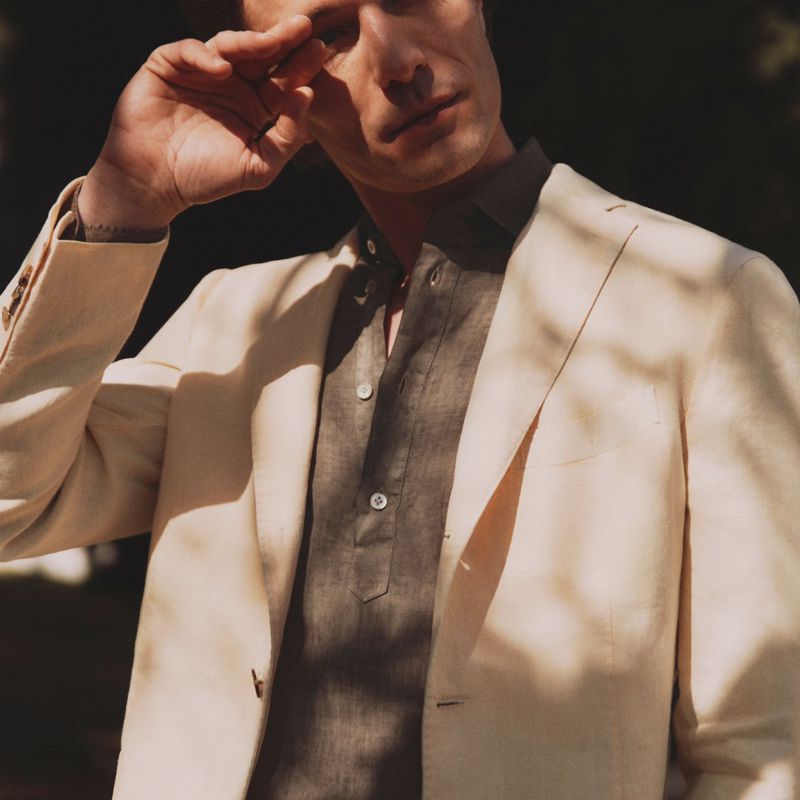 An adult male model in a neutral-toned blazer and casual shirt stands outdoors with shadows and dappled light across his face and outfit.