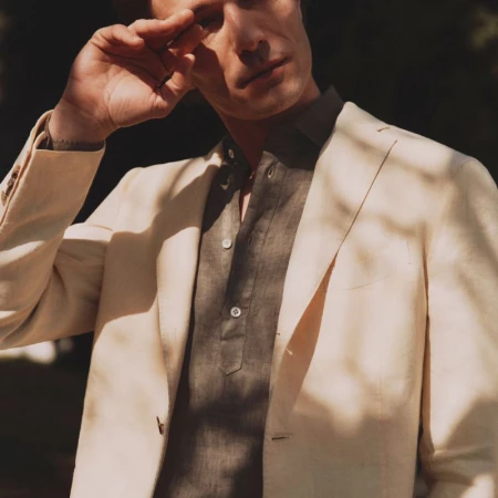 An adult male model in a neutral-toned blazer and casual shirt stands outdoors with shadows and dappled light across his face and outfit.