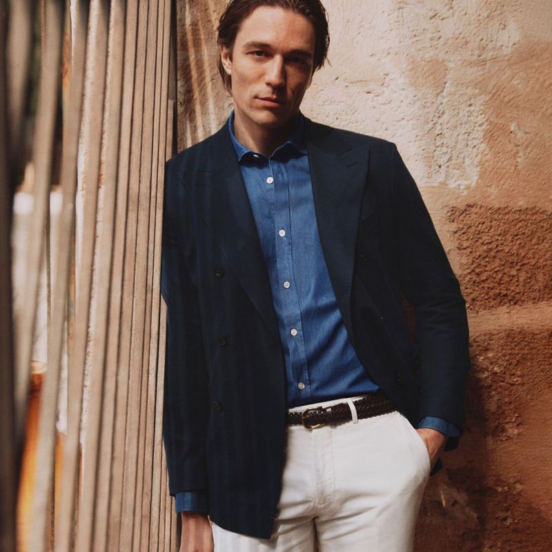 A stylish adult male in a blue shirt and navy blazer poses against a textured wall and wooden fence, exuding a casual yet refined look.