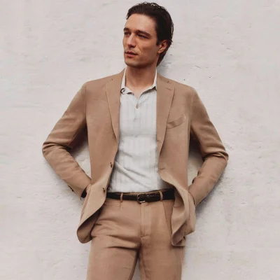 Man wearing a camel blazer and matching trousers with a white polo shirt against a textured white wall