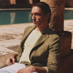 Male model in an olive green blazer and light yellow sweater seated near stone pillars outdoors